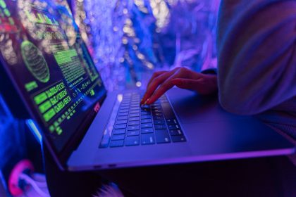Mãos digitando em um laptop com gráficos de cibersegurança iluminados por luz roxa.